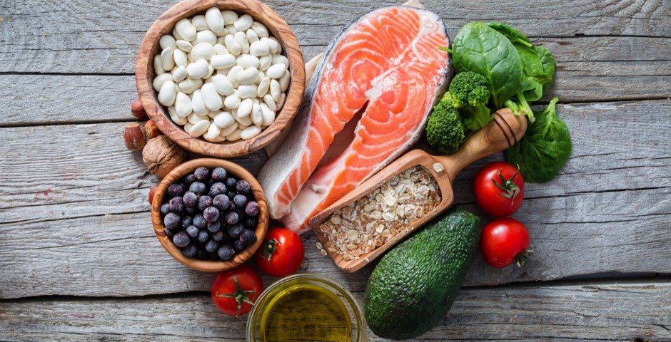 Olika livsmedel som lax, bönor, bär, grönsaker och olivolja upplagda på ett träbord.