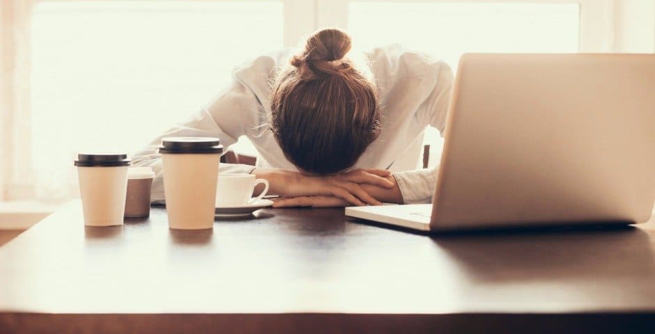 En kvinna ligger med huvudet i händer på ett bord med kaffekoppar och en dator.