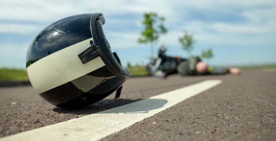 En motorcykel hjälm ligger på en väg och en person ligger längre fram vid en vält motorcykel.