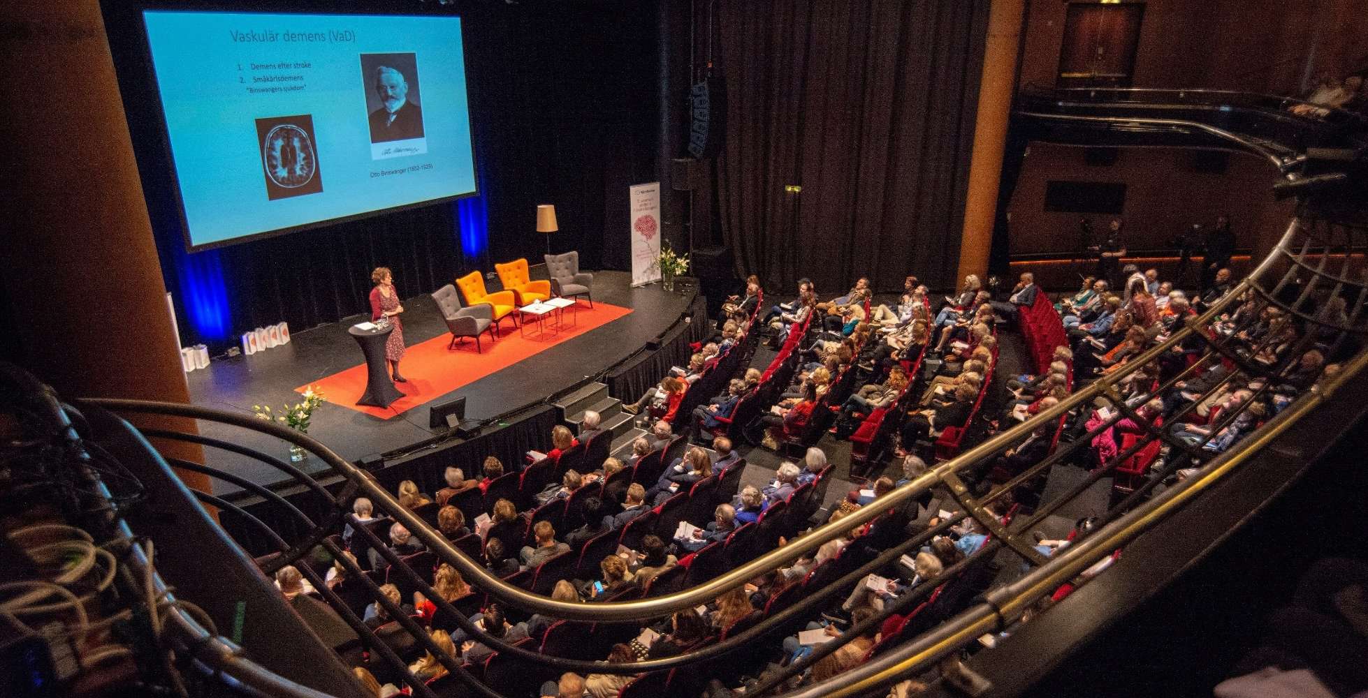 Publik i en sal som tittar mot en scen där en person står och talar.