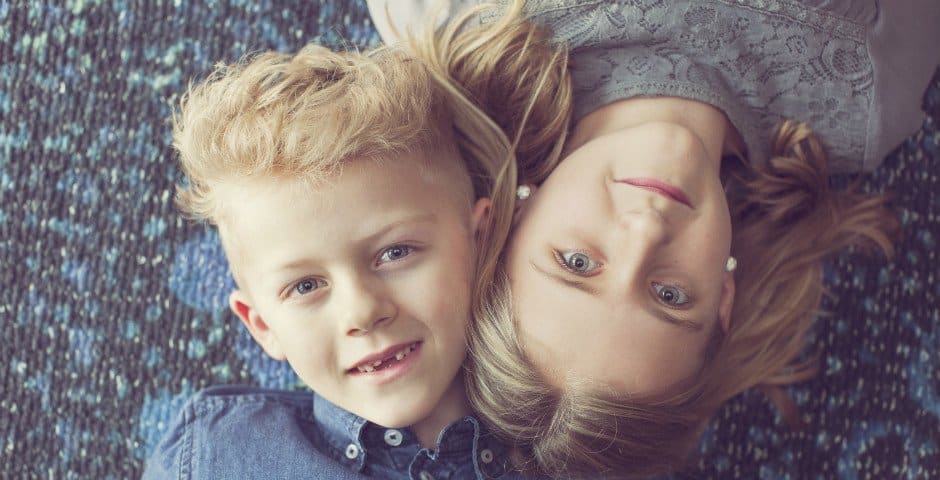 Två barn ligger på golvet med huvuden mot varandra och tittar rakt upp mot kameran.