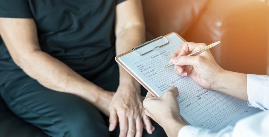 En hand som fyller i ett formulär under ett möte mellan vårdpersonal och en person som sitter på en soffa.