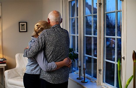 Ett äldre par kramar om varandra i sitt hem vid fönstret.