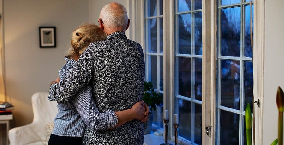 Ett äldre par kramar om varandra i sitt hem vid fönstret.