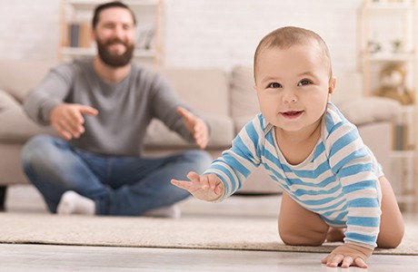 En bebis kryper på ett golv med sin pappa bakom sig.