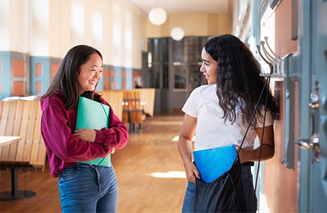 Två unga tjejer står i en skol-korridor och pratar.