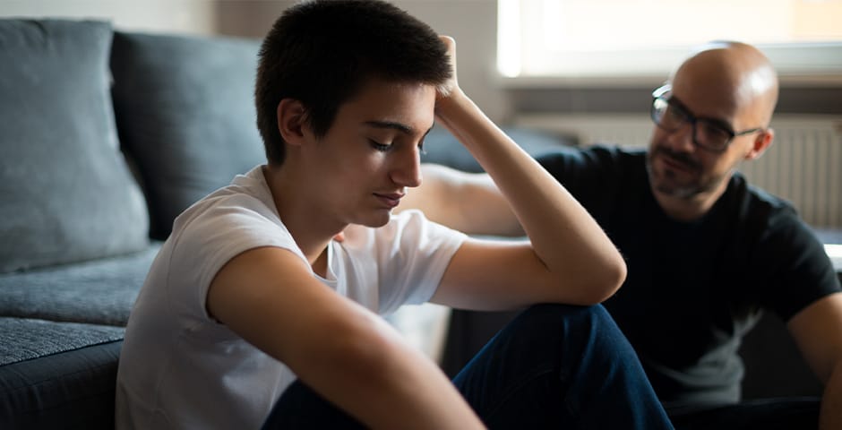 Ung kille sitter på golvet vid en soffa och hans pappa sitter bredvid med handen på axeln.